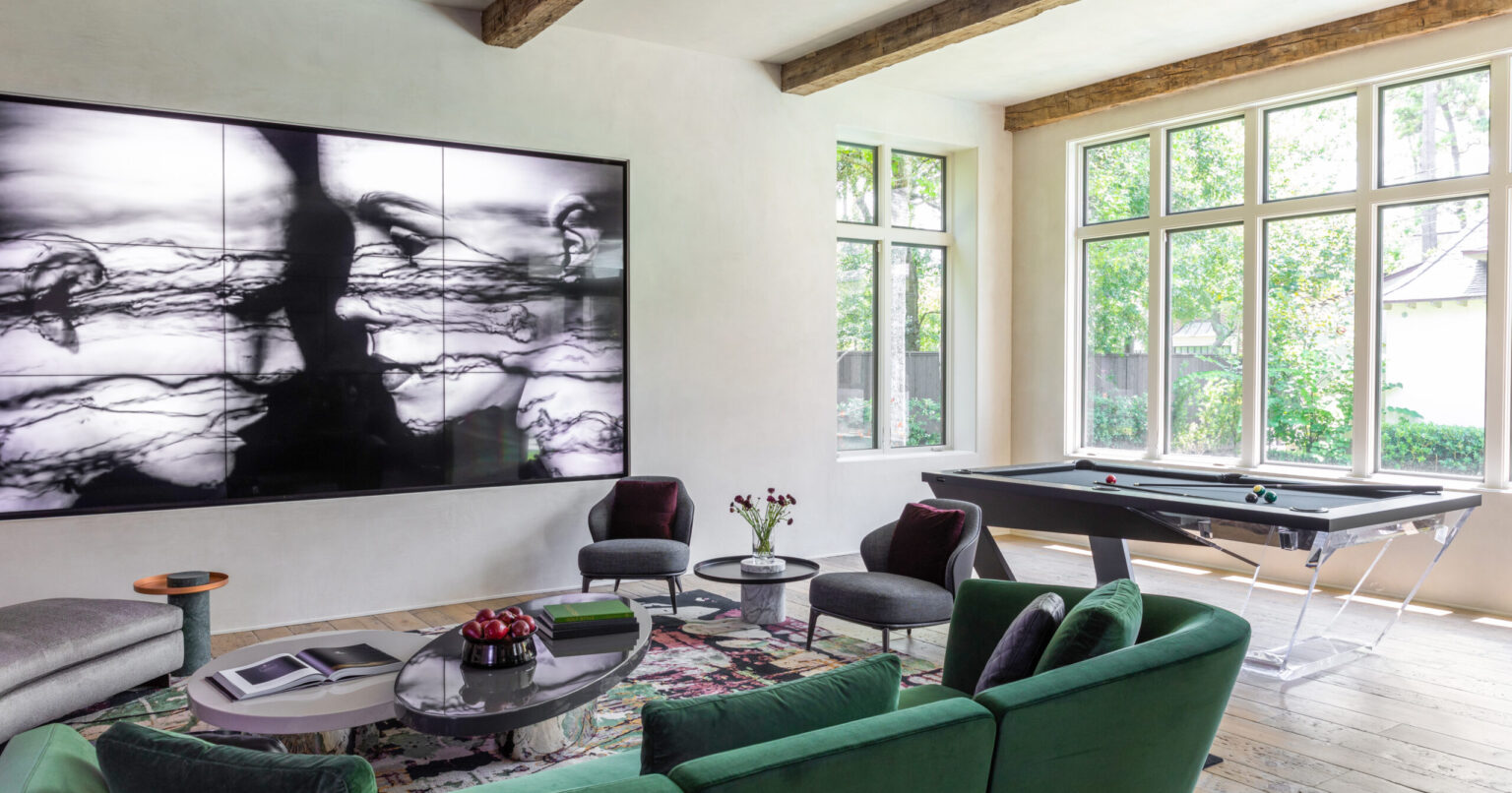 A modern living room highlights a large abstract art piece, green sofas, and a round coffee table with books and flowers. The Avettore Pool Table blends seamlessly under exposed wooden beams, while large windows let in natural light for a rustic touch.