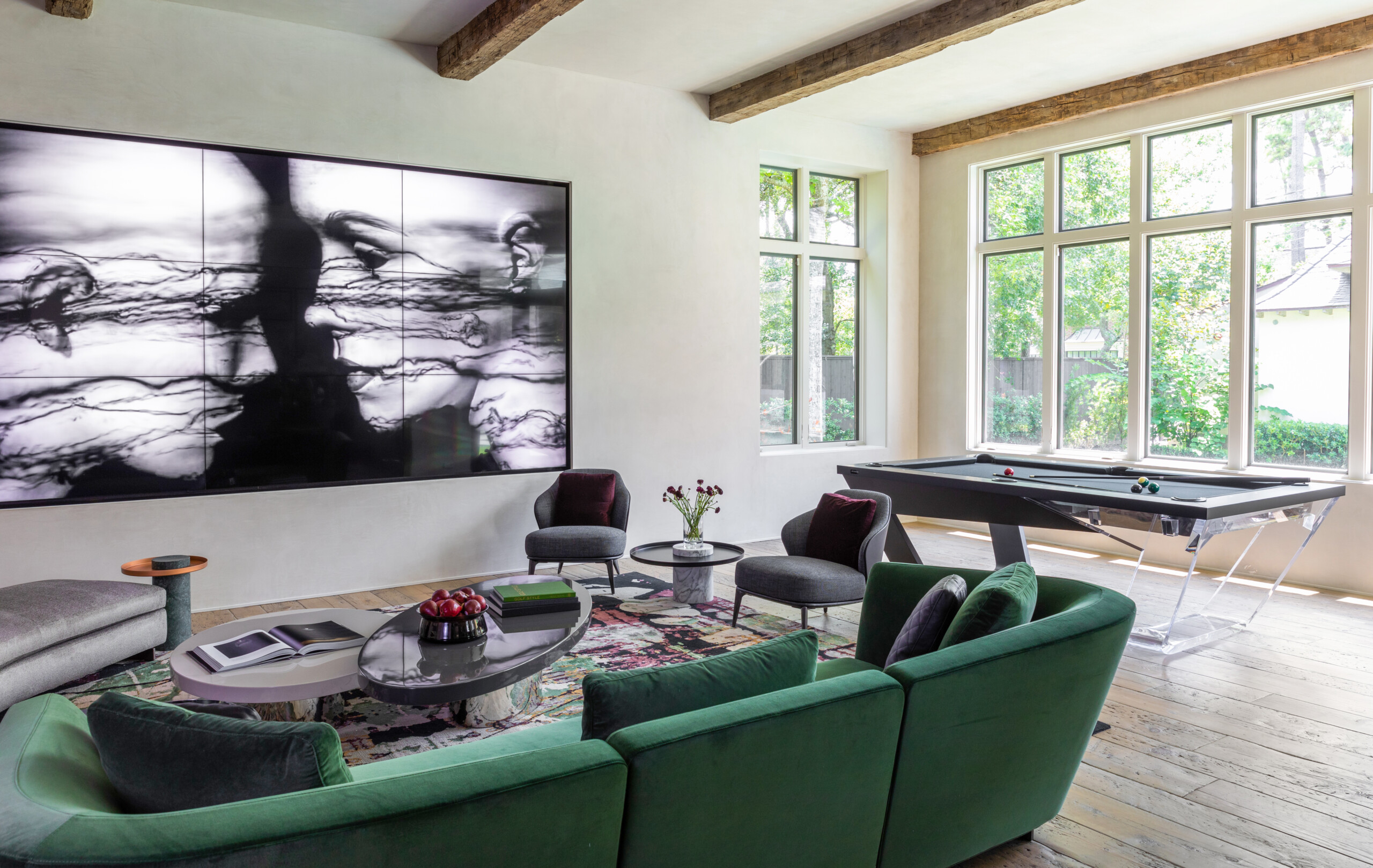 A modern living room highlights a large abstract art piece, green sofas, and a round coffee table with books and flowers. The Avettore Pool Table blends seamlessly under exposed wooden beams, while large windows let in natural light for a rustic touch.