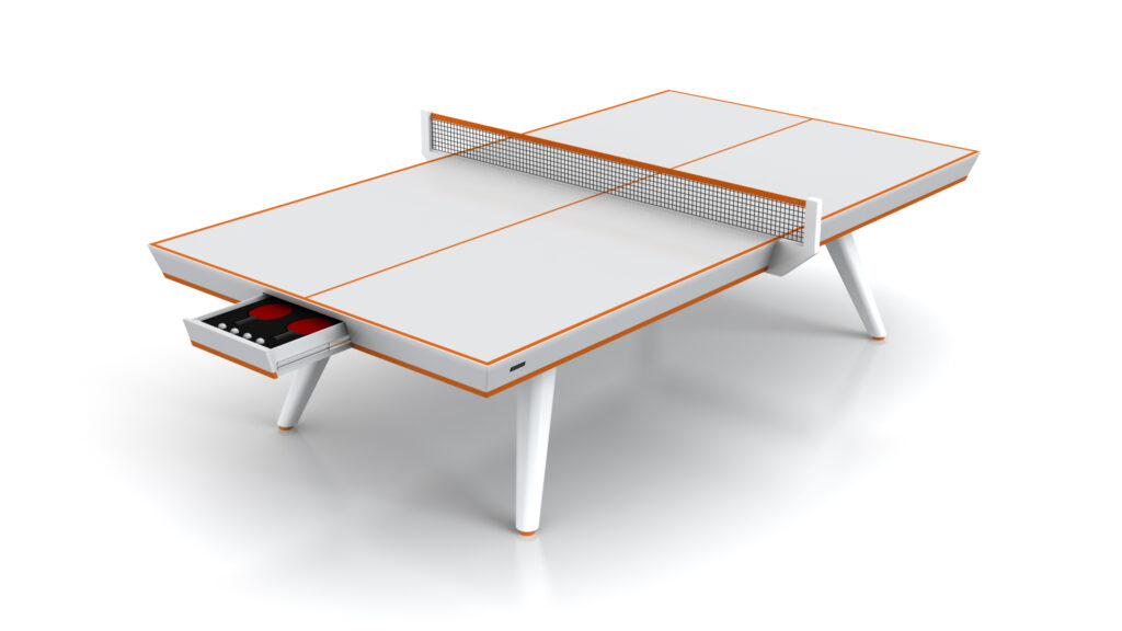 The Hamptons Ping Pong Table features a modern white finish with orange accents, angled legs, and a built-in drawer partially open to show table tennis paddles and balls inside.