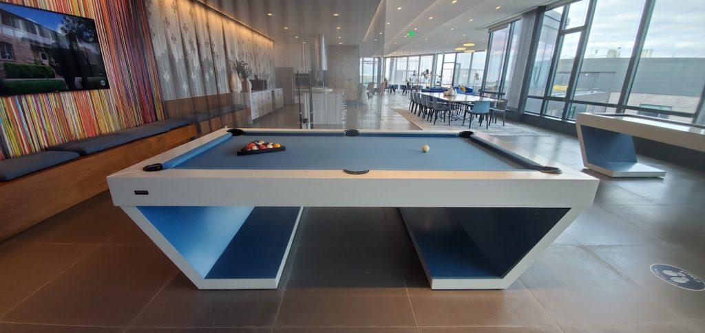 The modern office space showcases the luxurious Cabaret Pool Table with blue felt and a triangular rack of pool balls. It is brightly lit, features contemporary decor with colorful wall accents, and has a spacious open-plan layout with additional tables and chairs in the background.