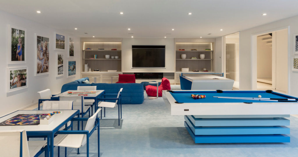 A modern entertainment room includes the luxury Arclight Pool Table and board games, complemented by a large TV and comfortable seating. White walls, adorned with framed pictures and recessed lighting, create a spacious, inviting atmosphere.
