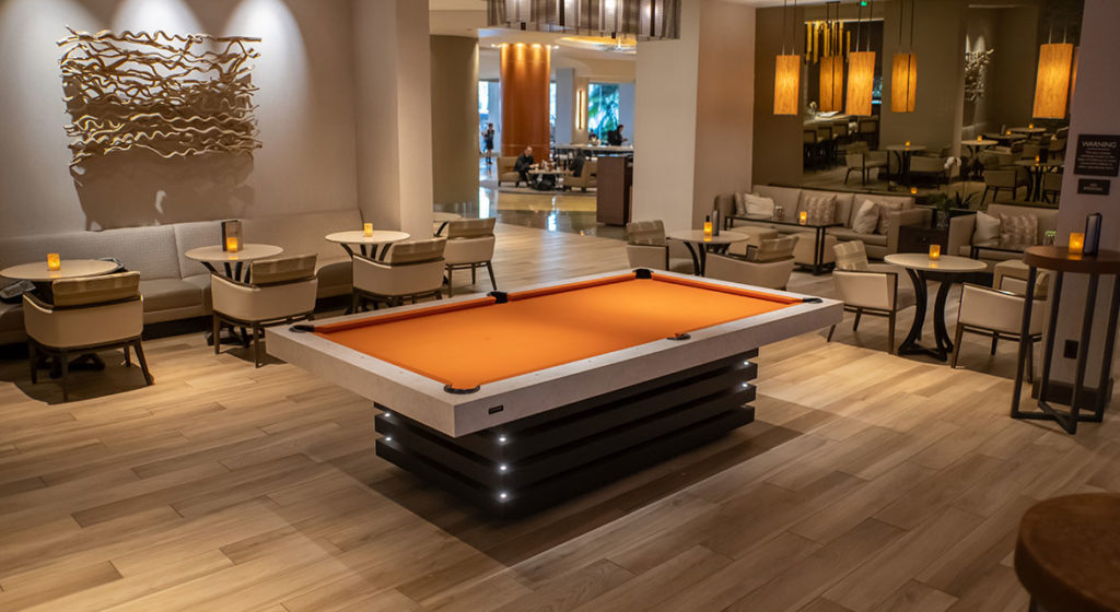 A modern lounge showcases a luxurious orange Arclight Pool Table on a light wood floor, surrounded by seating areas with beige chairs and small tables. Warm lighting and wall art enhance the cozy ambiance.