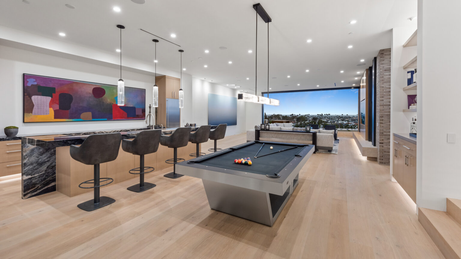 The open-plan living space showcases the Titan Pool Table as the centerpiece. A stylish bar with high stools and pendant lights is on the left, while a large window provides a scenic view. The artistic decor and light wood flooring enhance the luxurious ambiance.