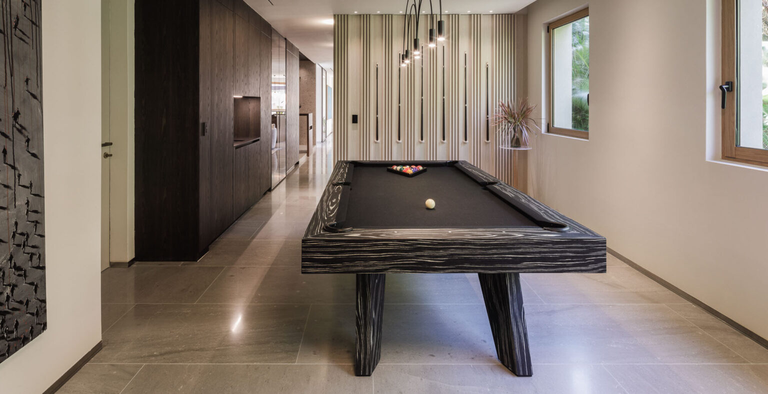 A modern interior showcases the Trigon Pool Table, epitomizing luxury with its sleek black design, a single white ball, and a triangle of colored balls. The room features polished stone floors, wood-paneled walls, contemporary lighting, and large windows that flood the space with natural light.
