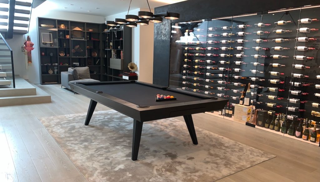 A modern room highlights luxury game tables featuring the Hamptons Pool Table in sleek black on a light gray rug. A wall-mounted wine rack displays various bottles, while shelves with decor, a staircase, a sofa, and a vintage gramophone complement the setting.