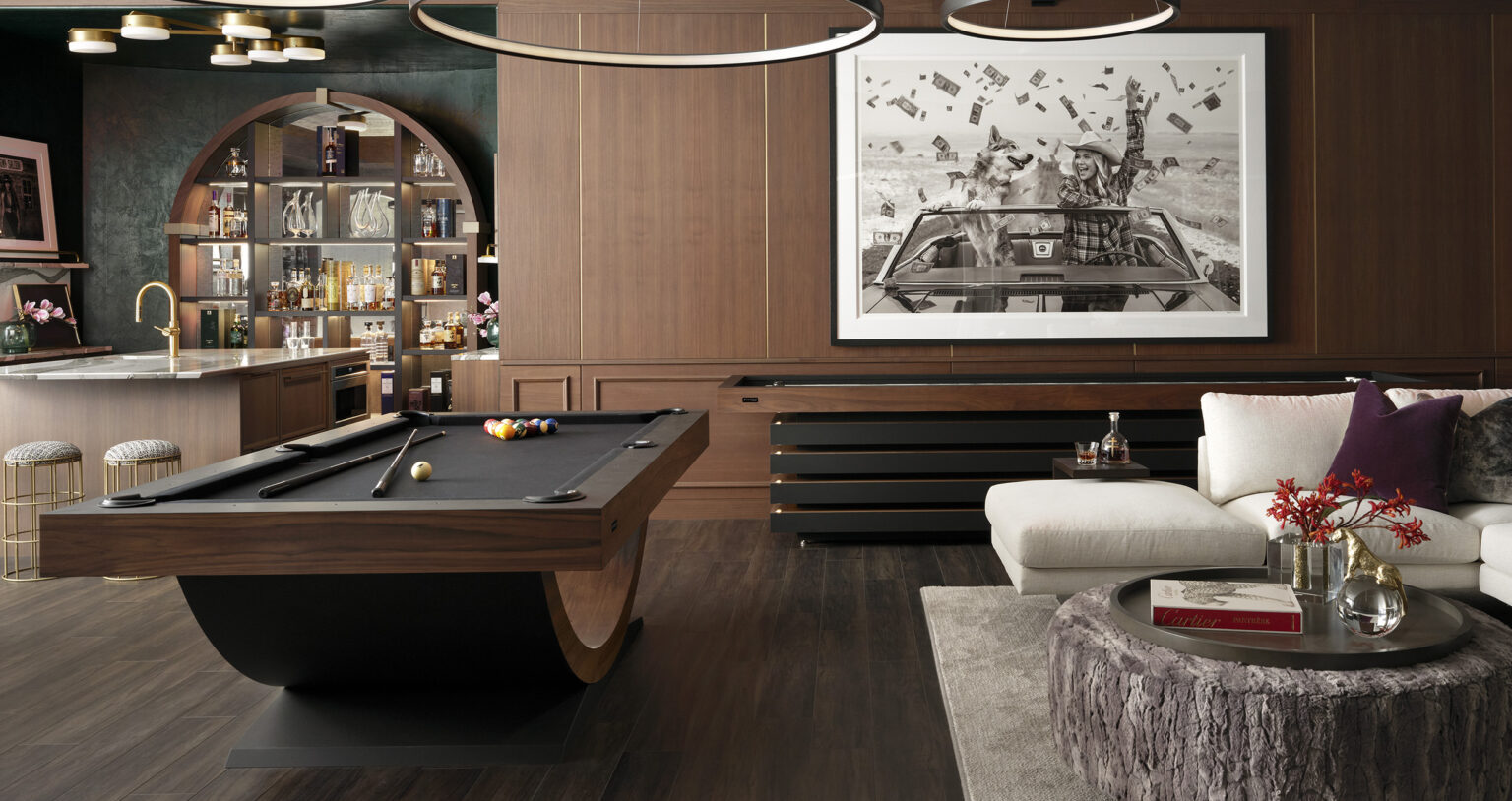 The chic game room showcases the Theseus Pool Table, accented by a bar stocked with liquor shelves. The lounge includes a white sofa and round wooden coffee table beneath a large black-and-white photograph on the wood-paneled wall.