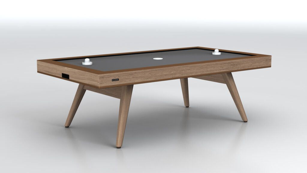 The Hamptons Air Hockey Table features a dark playing surface, angled wooden legs, two white strikers, and a central puck drop opening, all set against a plain white background.