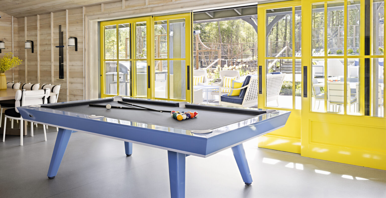 A modern room features the luxury Hamptons Pool Table, complete with cue sticks. Bright yellow-framed glass doors reveal an outdoor area with patio furniture. Sunlight streams in, highlighting the rooms light wood walls and gray floor.