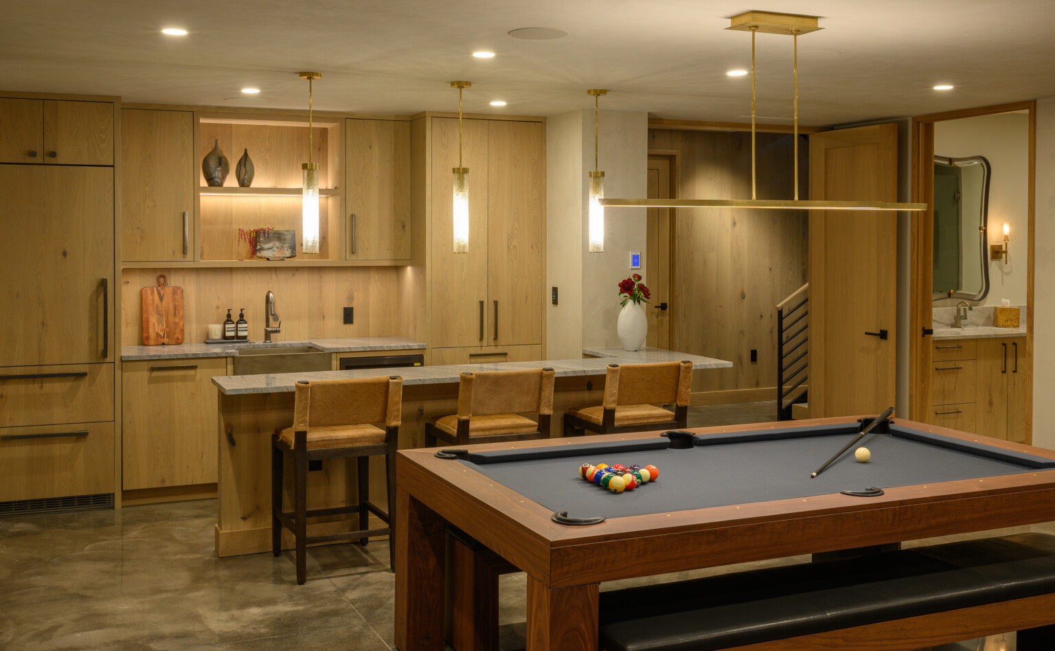 A modern game room features the luxurious Malibu Pool Table, with balls arranged and a cue stick resting nearby. A stylish home bar includes wooden cabinets, barstools, pendant lights, and decorative touches like vases and wall art.