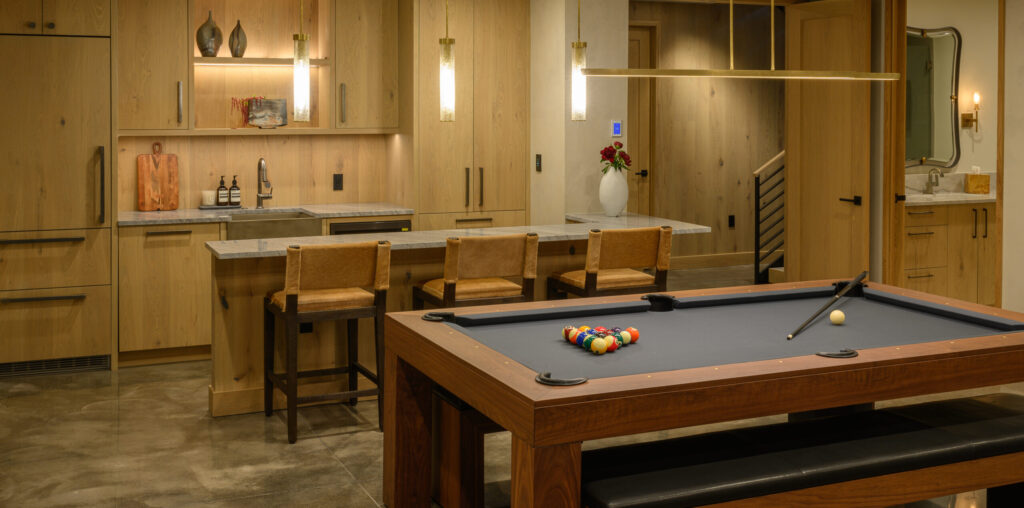 A modern game room features the luxurious Malibu Pool Table, with balls arranged and a cue stick resting nearby. A stylish home bar includes wooden cabinets, barstools, pendant lights, and decorative touches like vases and wall art.