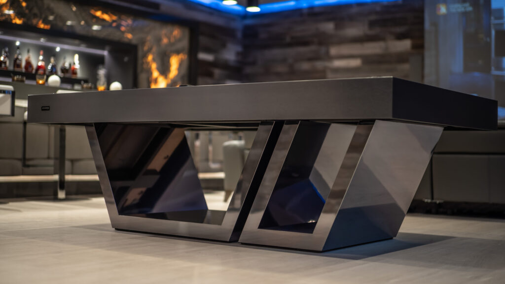 The Titan Pool Table features modern luxury with sleek metal angular legs. It sits elegantly in a stylish room, accented by shelves with bottles. Plush seating and blue ambient lighting further enhance the luxurious ambiance.