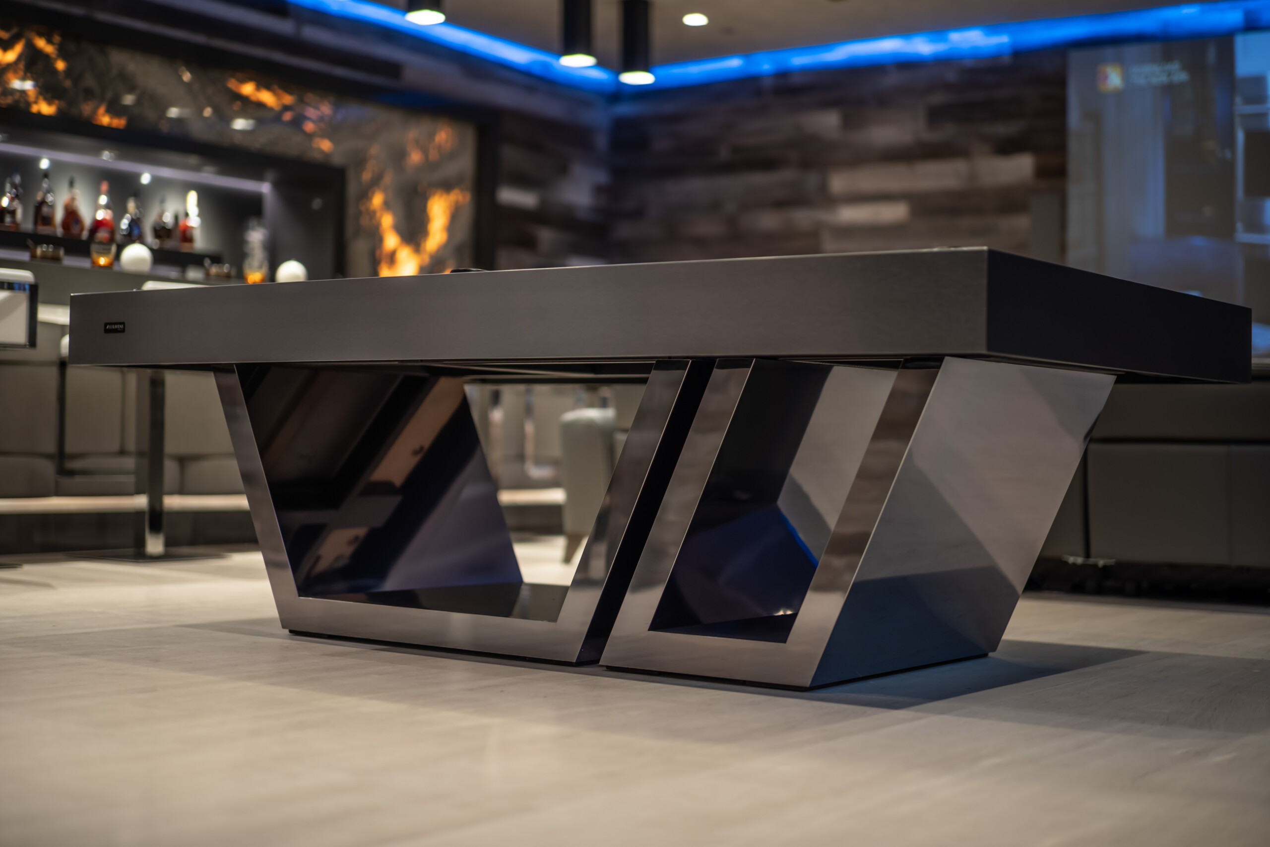 The Titan Pool Table features modern luxury with sleek metal angular legs. It sits elegantly in a stylish room, accented by shelves with bottles. Plush seating and blue ambient lighting further enhance the luxurious ambiance.