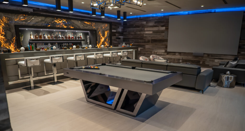 Contemporary entertainment room showcasing the elegant Titan Pool Table with modern geometric legs, a fully stocked bar with high stools, ambient lighting, and plush seating. The walls display a textured wood-like finish and feature a large flat-panel display.