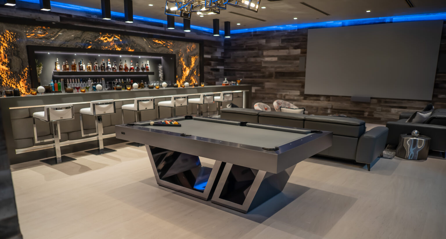 Contemporary entertainment room showcasing the elegant Titan Pool Table with modern geometric legs, a fully stocked bar with high stools, ambient lighting, and plush seating. The walls display a textured wood-like finish and feature a large flat-panel display.