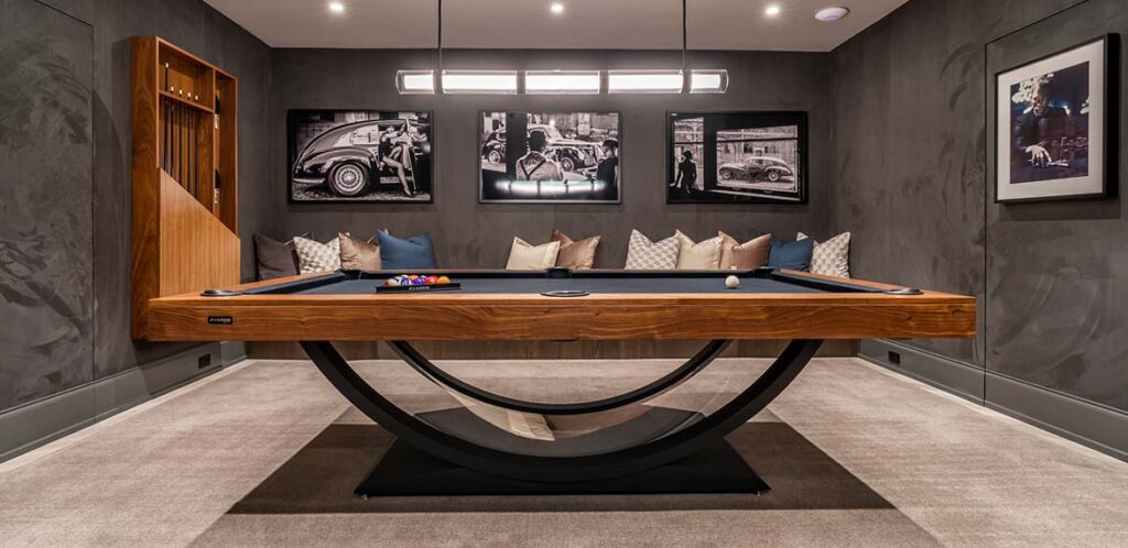 A modern game room features the luxurious Theseus Pool Table with black felt, decorative pillows on a cushioned bench, and framed black-and-white car prints on dark gray walls, all softly illuminated by overhead lighting.