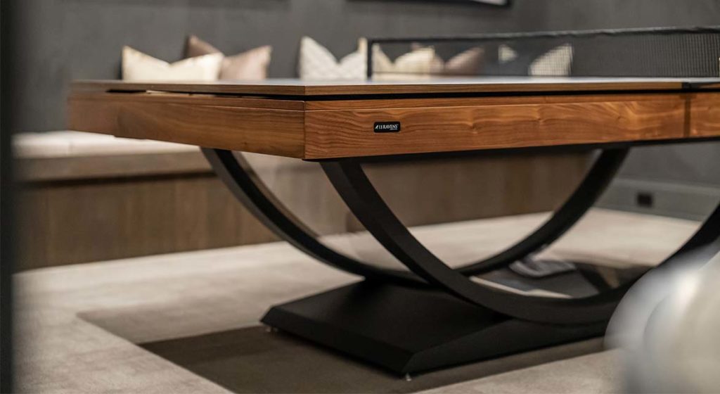 In a modern room, the elegant Theseus Pool Table features a wooden top and black curved metal legs. A plush bench with pillows enhances the settings luxury, while a small label adorns the tables side.