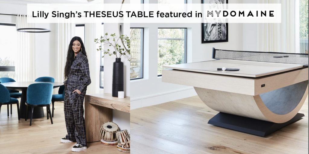 Lilly Singh’s Theseus Pool Table, featured in MYDOMAINE, stands out in a modern living room. The luxury table is surrounded by blue chairs, with large windows letting in natural light, as a woman in a patterned outfit poses nearby.
