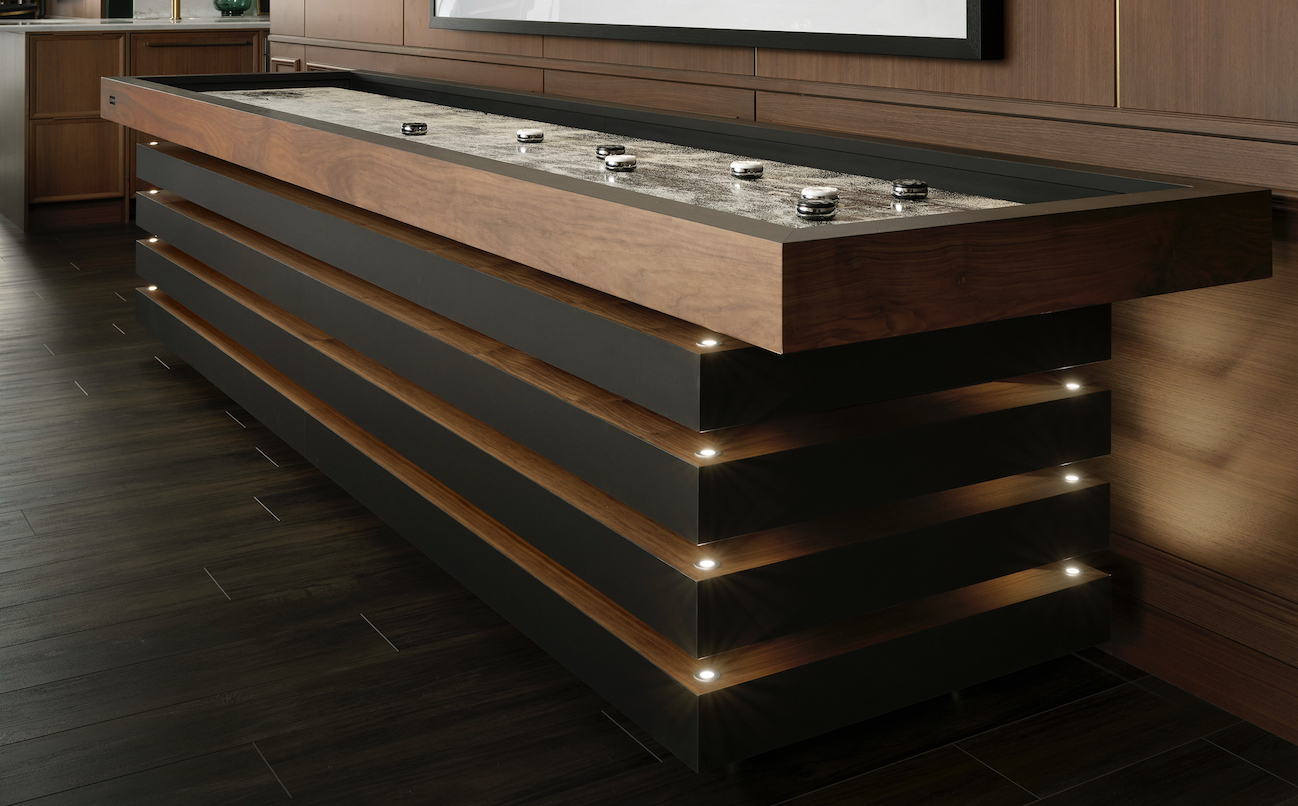 The Arclight Shuffleboard Table, featuring a wood and black metal design with integrated lighting, is showcased in a modern room with dark wood floors and paneled walls.