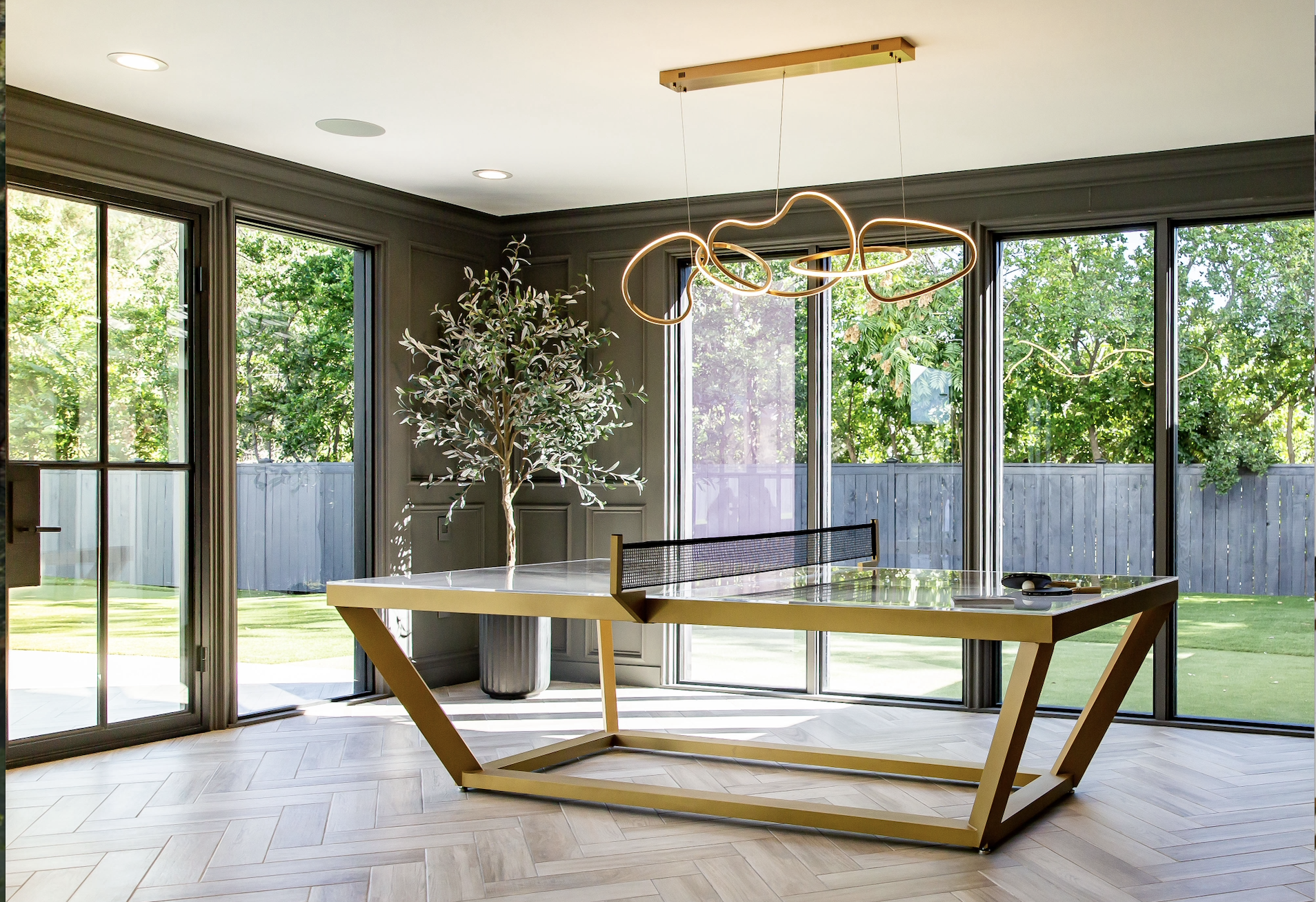 A modern room with large glass doors features a Vitro Ping Pong Table with a unique gold frame, herringbone wood floors, an abstract chandelier, and a tall potted plant; trees and a fenced yard are visible outside.