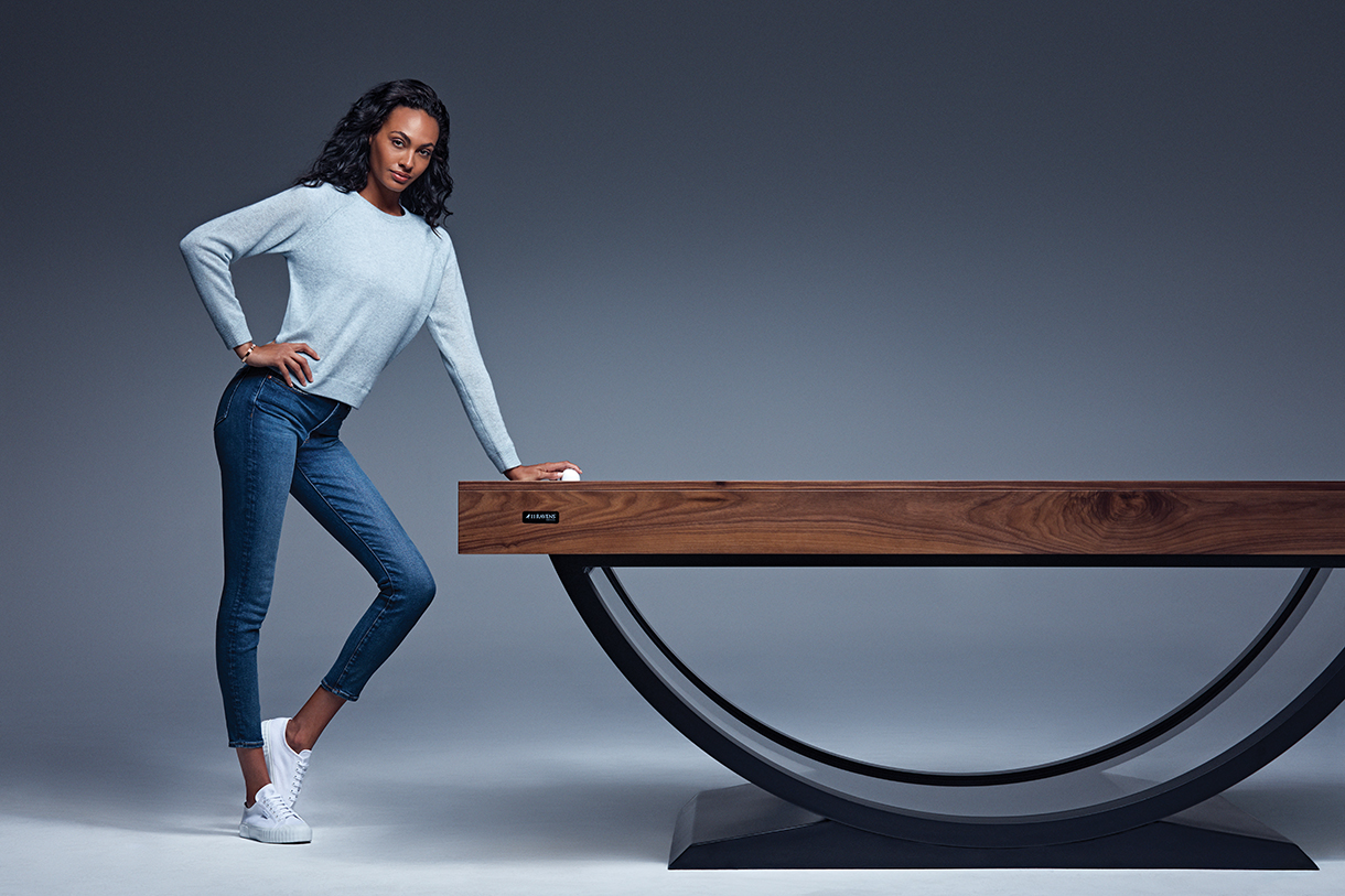 A woman in a light sweater, jeans, and white sneakers leans on a modern Theseus Air Hockey Table with a curved black metal base, set against a plain gray background.