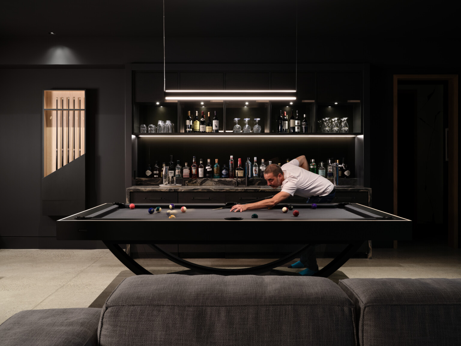 A person in a white shirt is playing pool on the sleek black Theseus Pool Table in a dimly lit room. The table stands out with a fully stocked bar and various bottles in the background. The modern decor with elegant dark tones creates an upscale atmosphere.