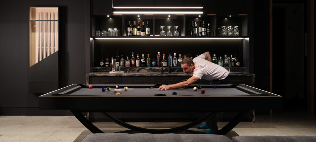 A person in a white shirt is playing pool on the sleek black Theseus Pool Table in a dimly lit room. The table stands out with a fully stocked bar and various bottles in the background. The modern decor with elegant dark tones creates an upscale atmosphere.