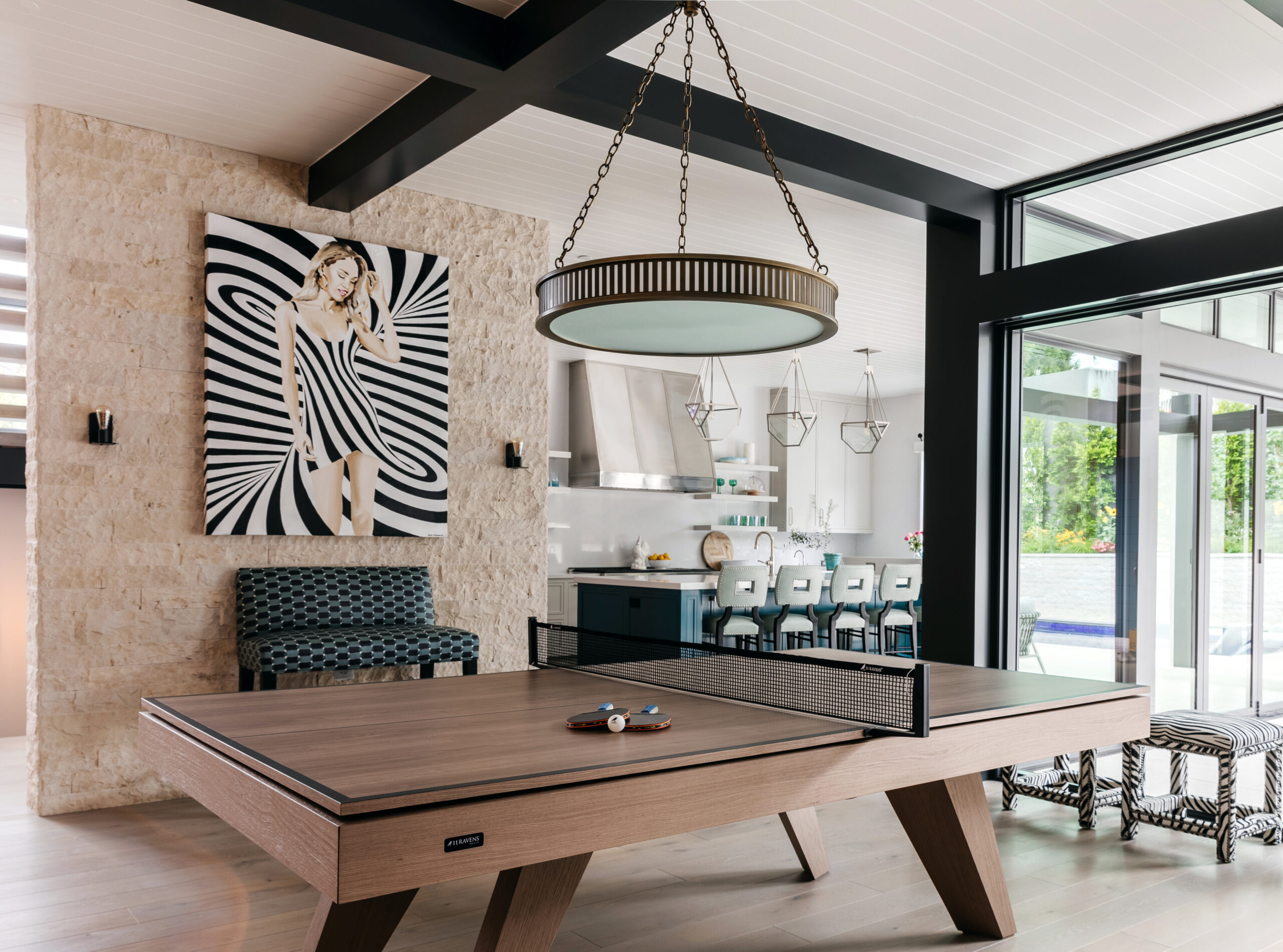 A modern room with a Trigon Ping Pong Table, a kitchen area with blue barstools, large windows, a round chandelier, and black-and-white artwork of a woman in a striped dress on a textured wall.