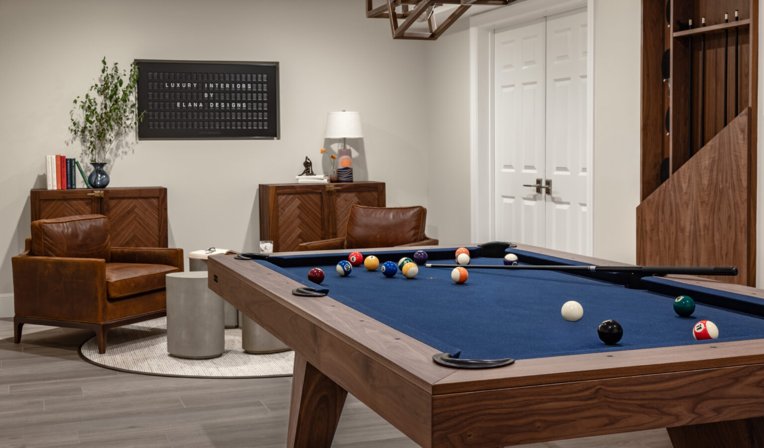 A stylish room features the luxurious Trigon Pool Table with a blue felt top, surrounded by brown chairs and a round rug. Shelving and a lamp sit in the background, while an elegant wall piece hangs above a wooden cabinet.