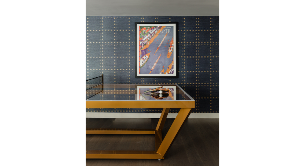 The stylish room, featuring the luxurious Vitro Ping Pong Table with its modern glass top and angular gold frame, exudes sophistication. A magazine cover showcases an aerial street scene against a dotted wall pattern, while the dark wood floor enhances the elegant ambiance of this high-end setting.