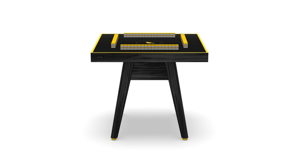 The Hamptons Mahjong Table is a modern black table with yellow accents, featuring a rectangular surface and two stacked rectangular blocks on top, displayed against a white background.