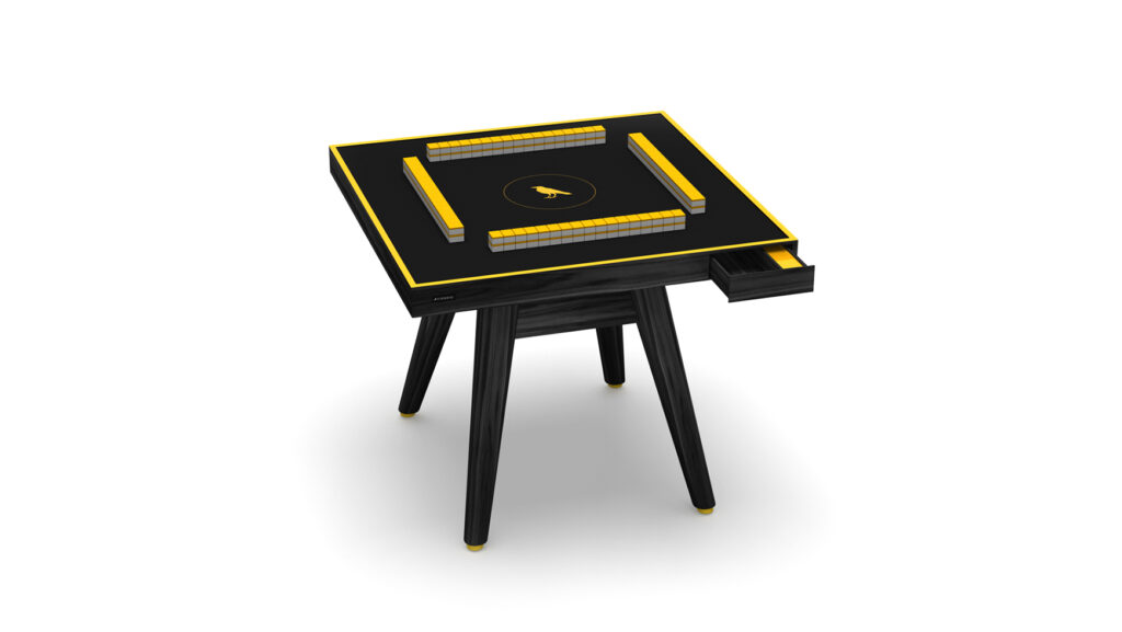 The Hamptons Mahjong Table is a modern black table with yellow accents, built-in drawers, and arranged mahjong tiles on top. It stands on four legs and is displayed against a white background.