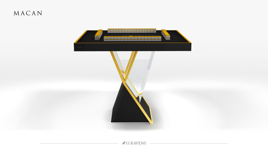 The Macan Mahjong Table showcases a modern black and gold design with a sleek glass and gold geometric base. Set with tiles, it’s displayed on a plain white background with "MACAN" in the upper left corner.