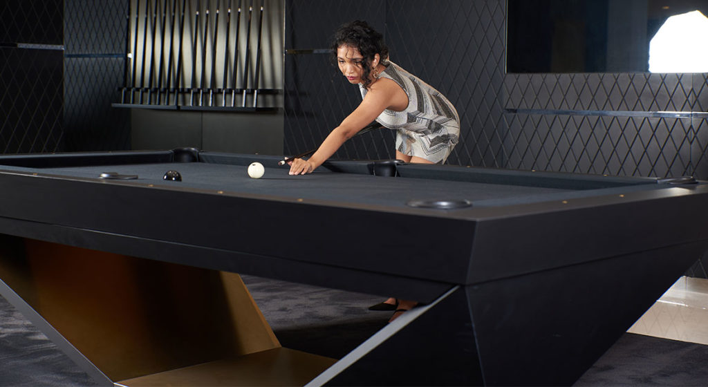 Understated lighting and a patterned wall frame the dark, elegant interior where someone plays on an R11 Stealth Pool Table. The table boasts a sleek, geometric design. With focus and precision, the player aims to strike the cue ball.