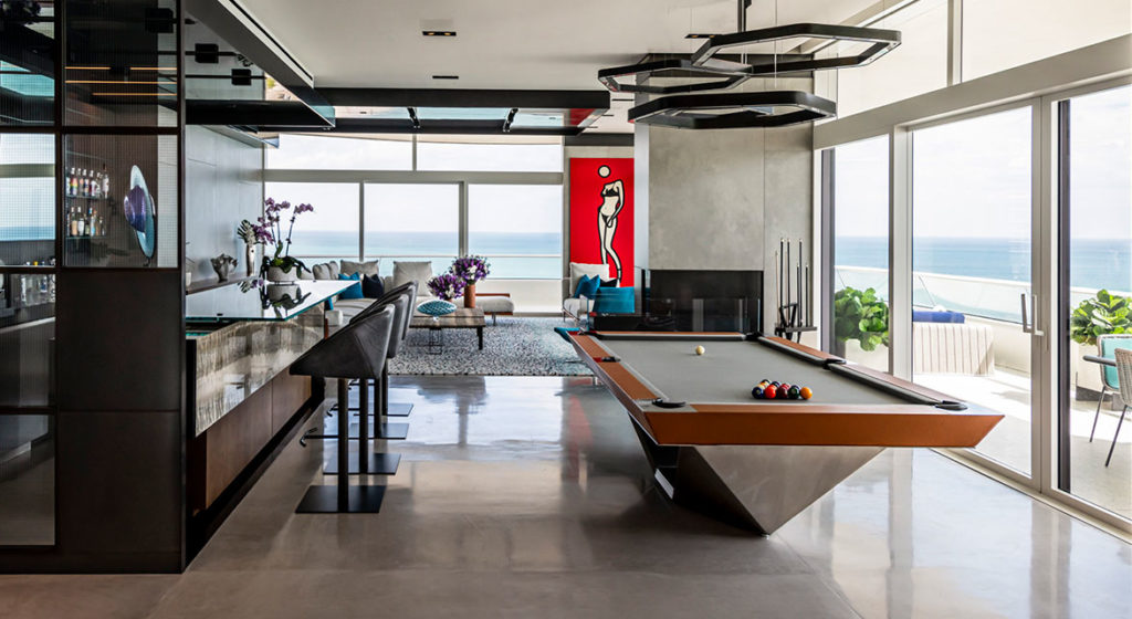A contemporary living space showcases the R11 Stealth Pool Table with a geometric base, a well-stocked bar, and a lounge adorned with vibrant red art. Large windows offer ocean views, enhancing the rooms open and stylish ambiance.