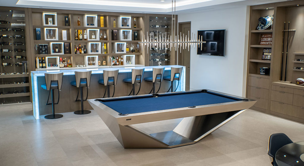A modern game room features the luxurious R11 Stealth Pool Table and a backlit bar with high chairs. Shelves display bottles and glasses, while a contemporary chandelier lights up the space. A helmet and sports memorabilia enhance the sleek decor.