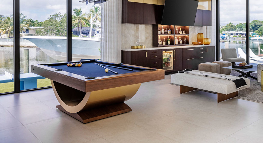 A modern living room features the luxurious Theseus Pool Table, large waterfront-view windows, a bar area with bottle shelves, a TV, and contemporary seating. Neutral tones with wooden and beige accents enhance the opulent ambiance.