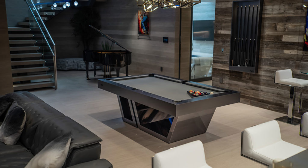 A stylish modern living room features a Titan Pool Table with a black surface and ball triangle. The space has a leather sofa, white chairs, piano, and unique chandelier. Wooden-textured walls add elegance while stairs lead upstairs.