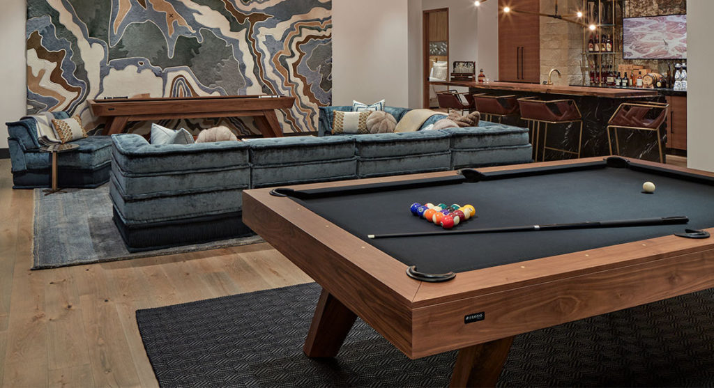 A stylish lounge features the luxury Trigon Pool Table with a black felt surface and pool balls in the foreground. A blue sectional sofa complements artistic wall decor, and a bar area with stools and bottles enhances the leisure ambiance.