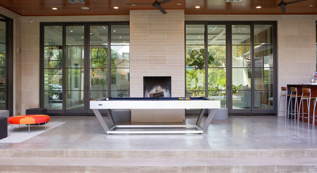 The modern room showcases a Vitro Pool Table positioned elegantly by the fireplace. Large glass doors open to lush greenery, while contemporary seating and a chic bar area enhance the sophisticated ambiance on the right.