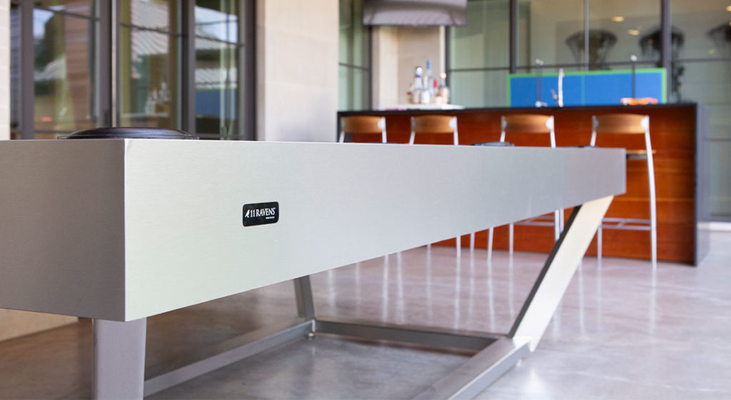 A metallic indoor shuffleboard table enhances the luxury of the Vitro Pool Table on polished concrete. A wooden bar and stools stand in the background against a stocked counter, while large windows flood the room with natural light.