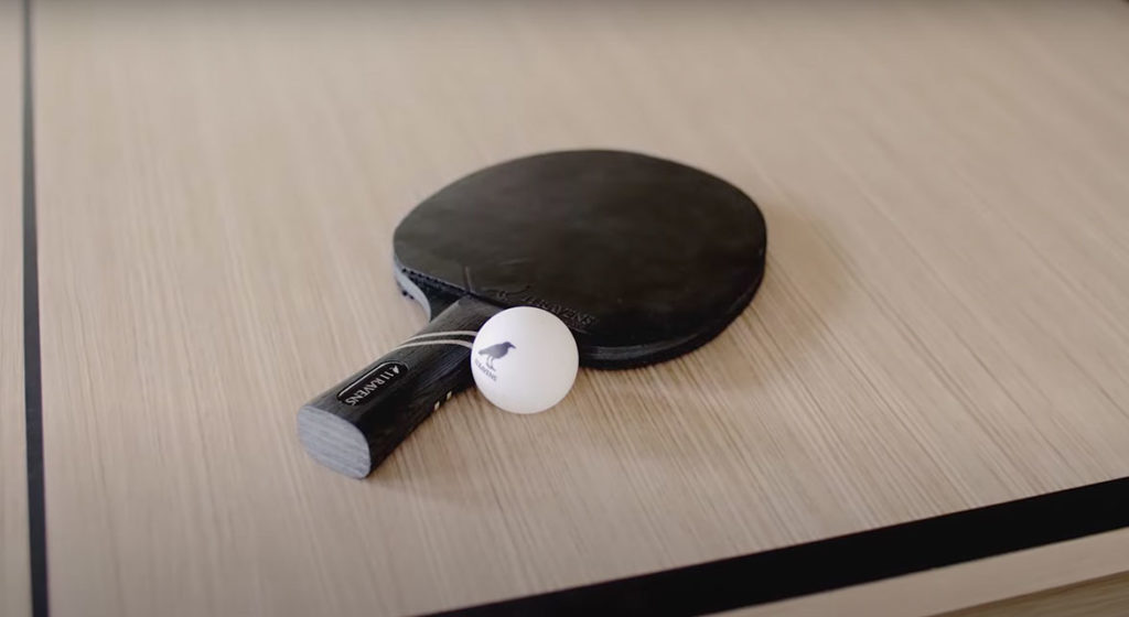 The 11R Racket, a luxury black table tennis paddle, and a white ping pong ball elegantly rest on a light wooden table with a sleek black edge line.