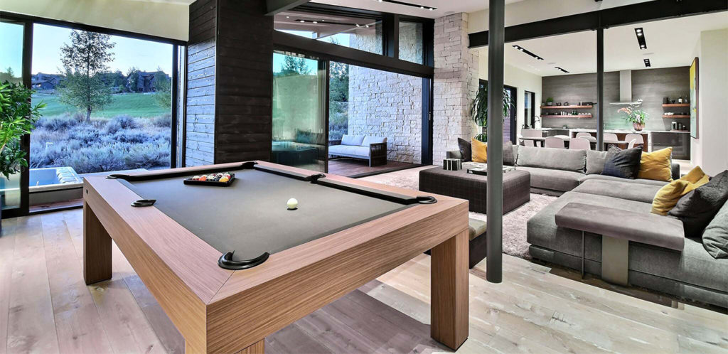 A modern living room showcases a black pool table from the recent work of 11 Ravens in the foreground. Large windows on the left offer scenic views of an outdoor landscape, featuring a spacious gray sectional sofa, a stone wall, and stylish decor.