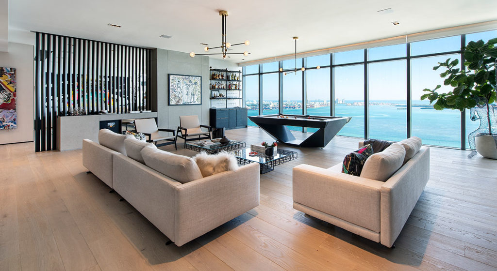A modern living room with ocean-view floor-to-ceiling windows includes beige sofas, a Stealth Pool Table - Quick Ship, a shelved bar, abstract art, and a large potted plant. The decor is contemporary with light wood flooring.