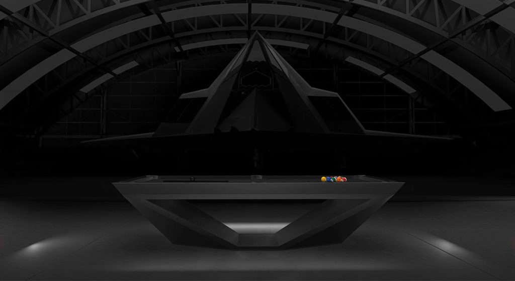 A sleek, modern Stealth Pool Table - Quick Ship with a triangular base stands in an industrial space. Colored billiard balls are arranged on it, while a large stealth aircraft in the background adds to the futuristic feel. Enhance your space swiftly and elegantly with quick ship options.