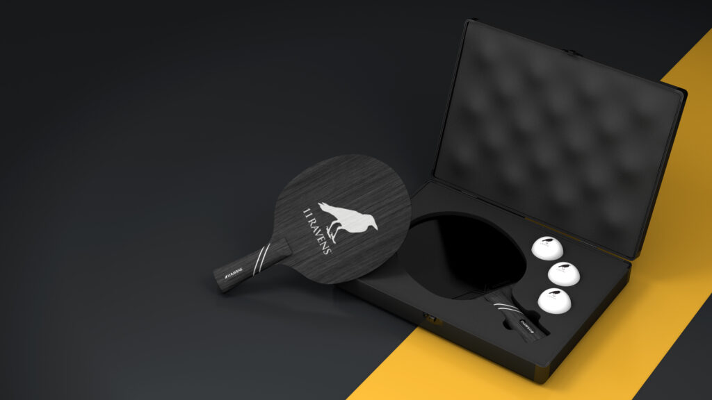A black paddle with a stylized bird logo and "RAVENS" in white, three white balls, and a Ping Pong Case with foam lining are shown on a black and yellow surface.