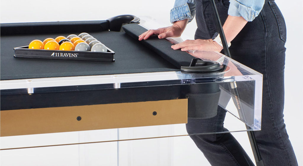 A person arranges billiard balls in a triangular rack on the Malibu Lucite Pool Table with black felt and transparent legs. The modern masterpiece gleams as the denim-clad player holds a cue stick, ready for the game.