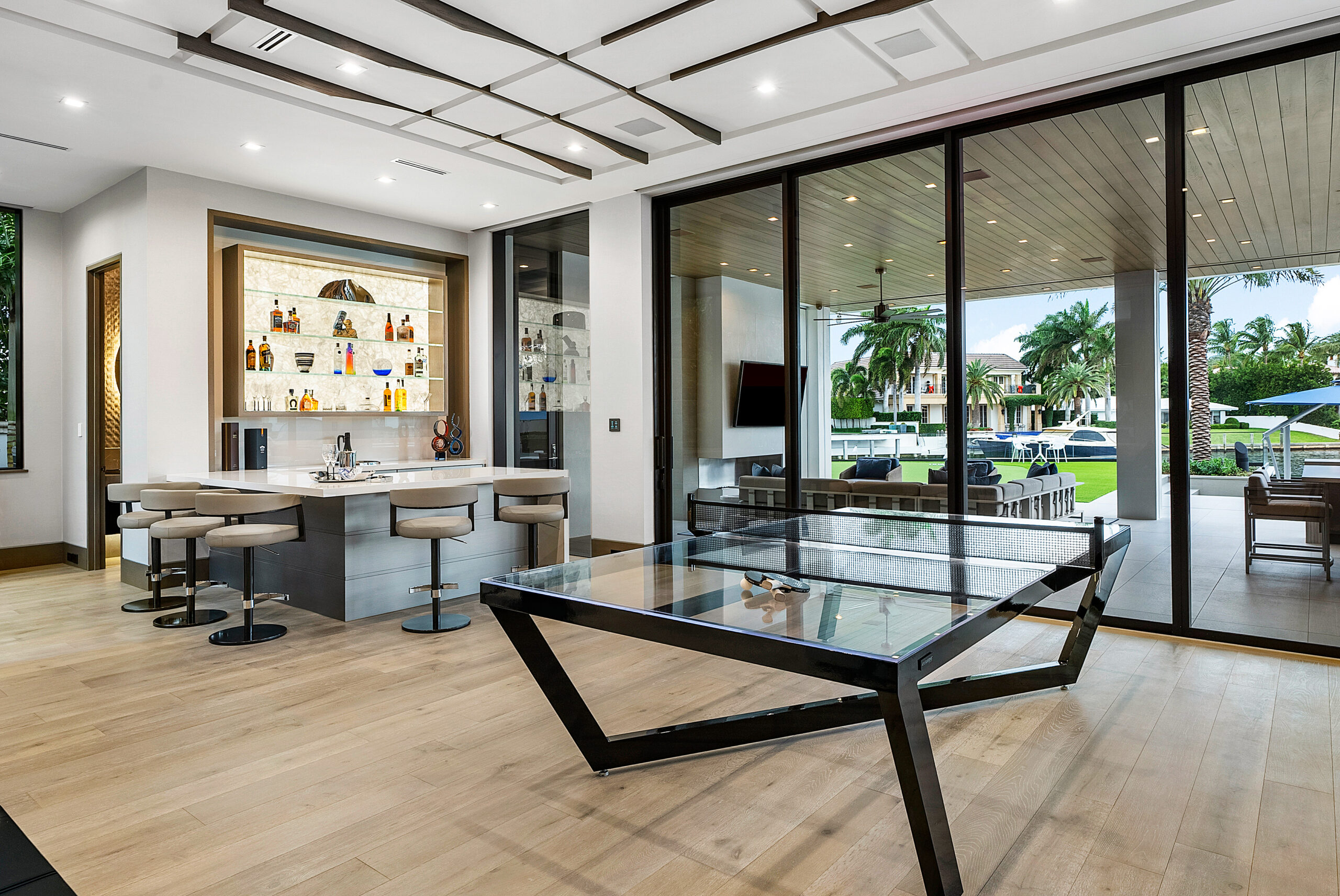 Modern home bar features light wood floors, three bar stools, illuminated bottle shelves, a Secavi Ping Pong Table with glass top, and large sliding doors leading to a green yard with palm trees.