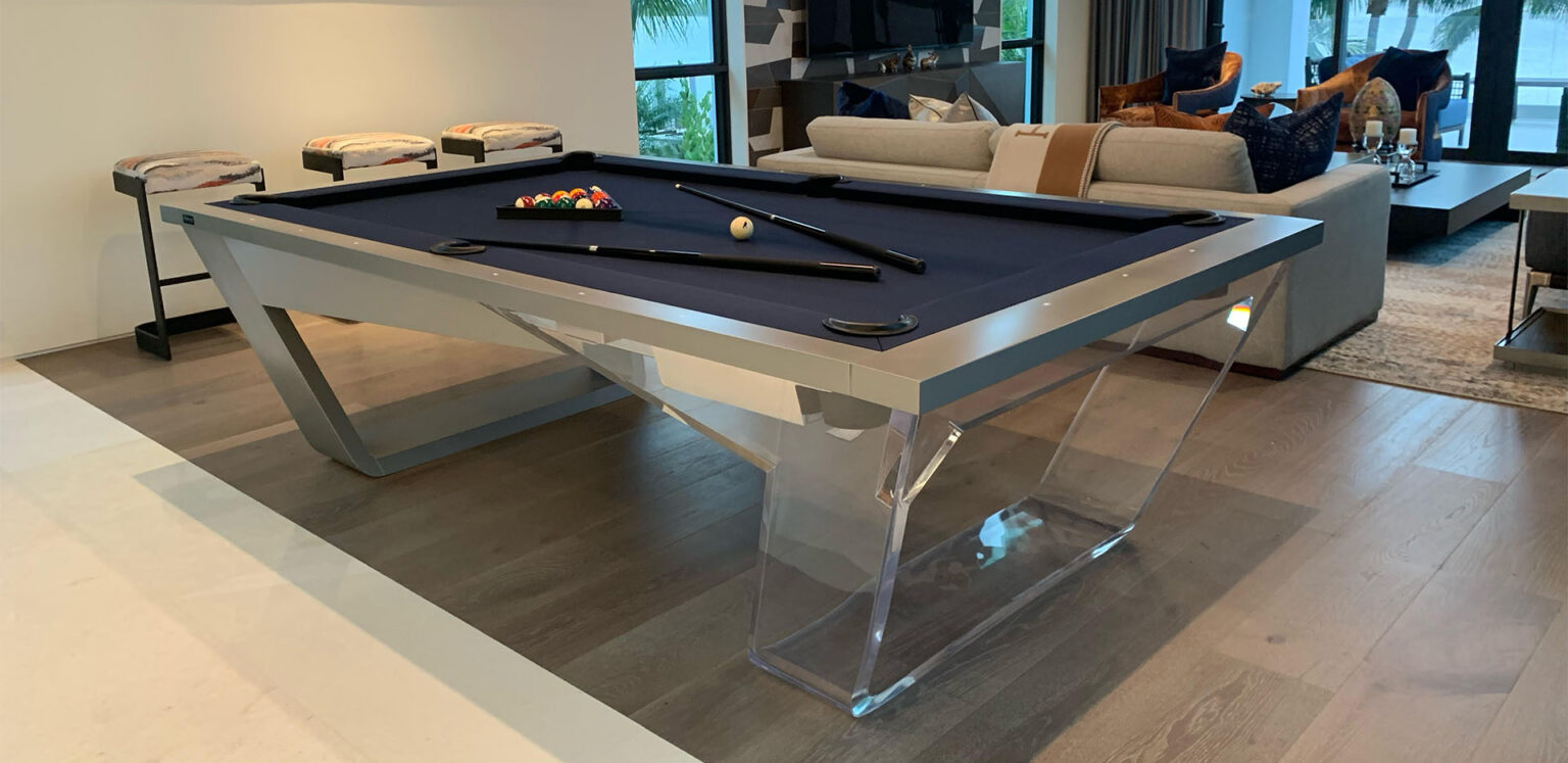 In a stylish living room, the Avettore Lucite Pool Table with its sleek design and transparent legs epitomizes luxury. Two cues and a triangle rack with balls rest on the table, complementing the rooms sofa, bar stools, and large window.
