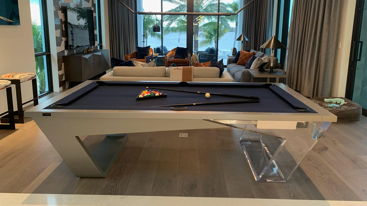 A modern living room features the Avettore Lucite Pool Table, elegantly adorned with a triangle rack of balls and a cue. The space is tastefully decorated with a sofa, pillows, and floor lamps, while large windows provide stunning views of palm trees and water.
