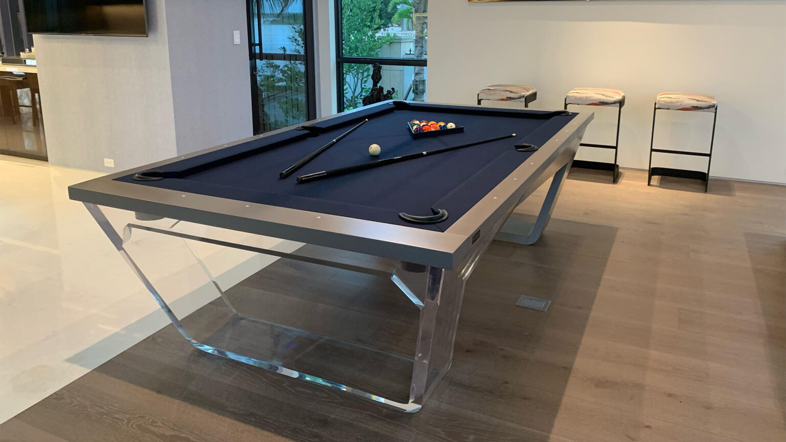 The Avettore Lucite Pool Table, featuring black felt and clear acrylic legs, is showcased in a stylish room. Two pool cues rest beside a racked triangle of balls on the table, with three stools nearby and large windows allowing natural light to pour in.